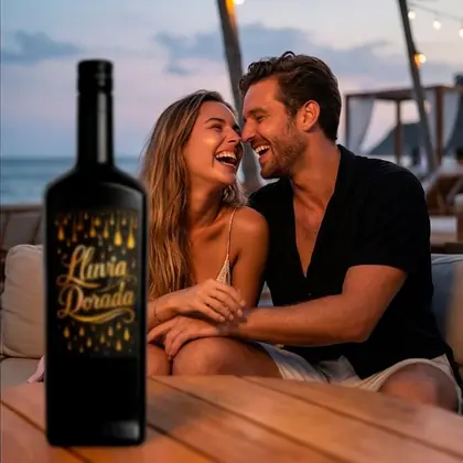 Pareja riendo en una terraza junto al mar al anochecer, con una botella de Lluvia Dorada en primer plano
